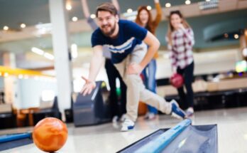 A young man sending a bowling ball down a lane while his friends watch and cheer in the background.