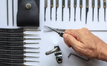 A man's hand holding a lock pick over an organized collection of other lock picks, a practice lock, and a carrying case.
