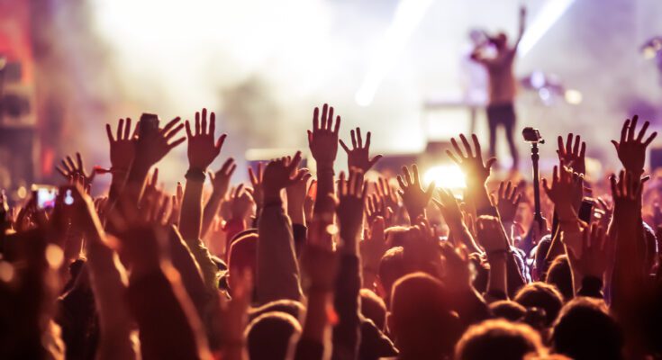 A crowd at a concert standing in front of a stage with their hands in the air. You can see a performer’s silhouette with a mic.