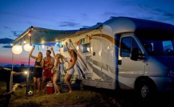 A young group of friends dancing, playing guitar, and celebrating in front of their RV decorated with lights and flags.