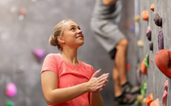 A young woman in athletic apparel stands at the bottom of an indoor rock climbing wall, looking up and rubbing her hands.