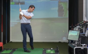 A young, well-dressed man in the middle of a golf swing in front of a golf simulator in his home.