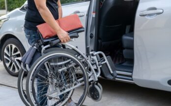 A person is folding a portable wheelchair and standing in front of a silver van with the side door open.