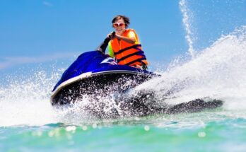 A teenager rides on a blue jet ski with a bright orange vest and water splashing everywhere in front of and behind them.