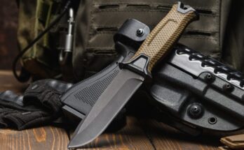 A combat knife sitting next to a military vest and a hand gun on a table. The combat knife is open and freshly sharpened.