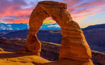 A gorgeous view of Delicate Arch in Arches National Park, Utah, at sunset. The sky is pink, blue, and purple.