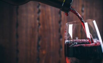 A close-up view shows a bottle pouring red wine into a wine glass. The background is brown and blurry.