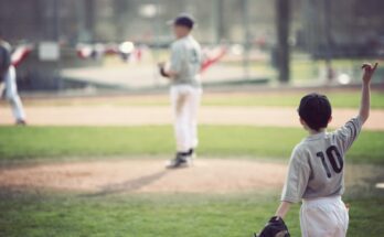 A community baseball game with young players in gray uniforms. We see a pitcher on the mound and outfielders.