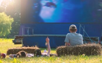 Three people are watching a movie in an open field. Two people lie in the grass, and one sits against a hay bale.