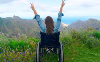 A wheelchair user experiencing international travel. She is throwing up her hands with peace signs looking over the mountain.