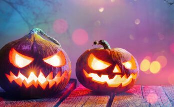 Two Jack O' Lanterns glowing with scary smiley faces sitting on a wooden patio deck. It's dark and eerie outside.