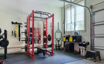 Home garage full of gym equipment including weights and bars, with two large windows and a white garage door.