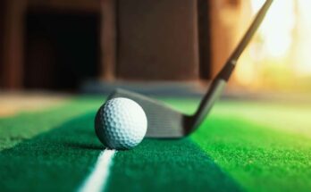 A close-up view shows a golf ball sitting on a manufactured putting green. A golf club appears behind the ball.