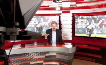 A TV anchorman sits in front of two TVS displaying two fighting athletes. A teleprompter is in the foreground.