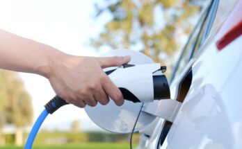 A close-up view shows a person plugging an electric charger into their white electric vehicle. The background is blurry.