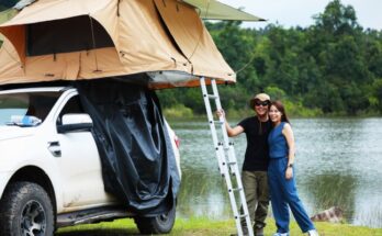 A man and a woman stand in front of the water, smiling. A rooftop tent sits on their white vehicle next to the water.