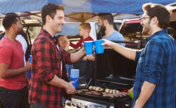 Five men, one little boy, and a woman are hanging out under a canopy tent, grilling behind a truck in a parking lot.