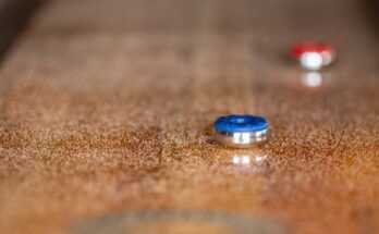 Close-up of a golden brown shuffleboard table with sand scattered across its surface. One blue puck and two red ones are visible.