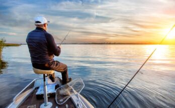 A man in a blue jacket and white baseball cap sits on the front of a fishing boat holding a fishing pole. The sun is setting.