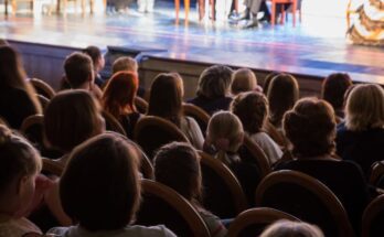 An attentive audience watches a live theatre performance. Actors sit around a table on stage in the distance.