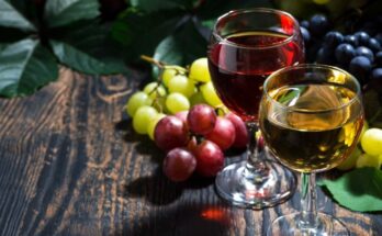 Two wine glasses sit on a wooden surface next to red and green grapes. One glass contains red wine, and the other white wine.