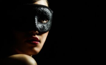 A woman in a black masquerade mask against a black background. Half of her face is covered by a shadow.