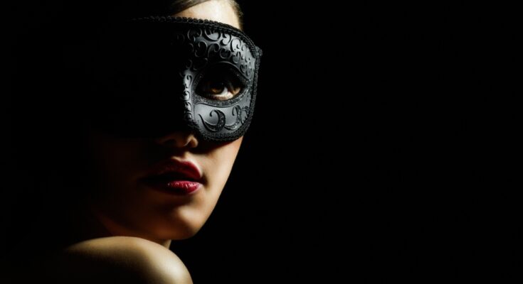 A woman in a black masquerade mask against a black background. Half of her face is covered by a shadow.