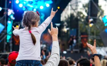 The back of a little girl with braids sitting on a man's shoulders both with "rock-on" hands at a concert during the day.