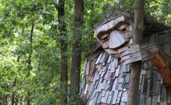 A troll made from wooden shingles in a forest, grabbing onto a tree with one hand and casting its eyes downward.