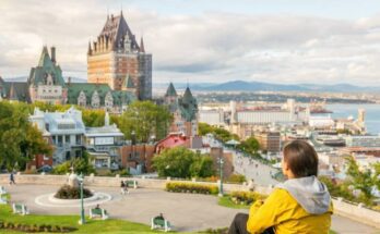A woman in a yellow jacket sits with her knees to her chest on a grassy knoll and overlooks Château Frontenac in Quebec City.
