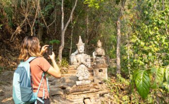 A woman wearing a backpack and using her phone to take a picture of a statue. The statue is sitting on a brick platform.