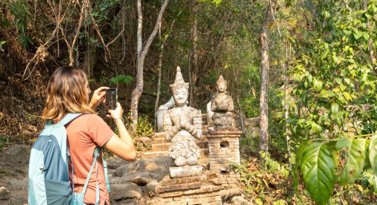 A woman wearing a backpack and using her phone to take a picture of a statue. The statue is sitting on a brick platform.