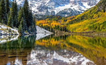 A crystal clear lake sits in a valley surrounded by tall, mature trees. A snowy mountain is in the background.