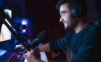 A young male live streamer in a dark room in the middle of a stream. There's equipment near him and he's smiling.