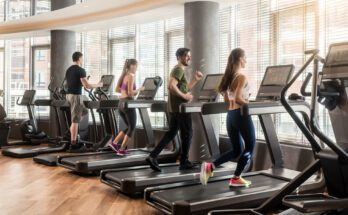 A row of treadmills placed in front of windows at the gym with users actively running and walking on the machines.