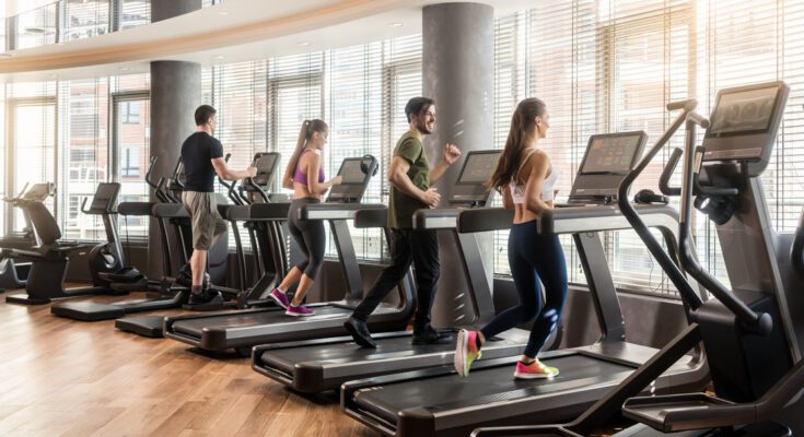 A row of treadmills placed in front of windows at the gym with users actively running and walking on the machines.