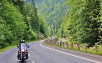 Two people ride on a large black motorcycle down a winding mountain road lined with a forest of green trees.