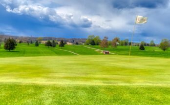 A white flag waves in the wind as it sits in the hole on a golf course’s putting green. Green grass surrounds it.