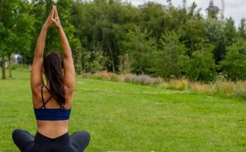 A woman sits cross-legged on a purple yoga mat with her arms raised above her head. The mat sits in the grass.