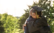 A young man wearing a black cap and gown for graduation is hugging an adult wearing a black suit. Trees are behind them.