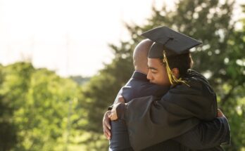 A young man wearing a black cap and gown for graduation is hugging an adult wearing a black suit. Trees are behind them.