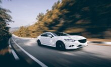 A white car with black tire rims speeds down a road with rows of trees blurring past it during the daytime.