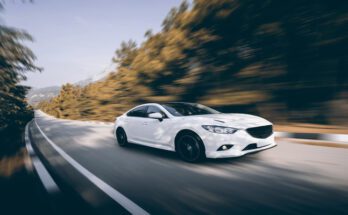 A white car with black tire rims speeds down a road with rows of trees blurring past it during the daytime.