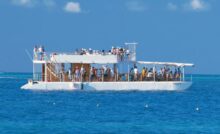 A party boat with an upper deck out on the water. Dozens of guests are mingling and talking on both levels.