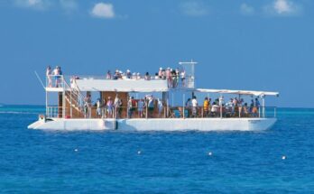 A party boat with an upper deck out on the water. Dozens of guests are mingling and talking on both levels.