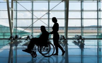 A silhouette of a female worker pushing a man in a wheelchair through an airport terminal with the runway in the background.