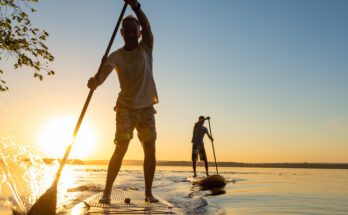 Two guys standing on a paddleboard with their paddles in the water. The water is calm and the sun is setting behind them.
