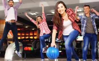 A woman in a plaid shirt rolls throws a blue bowling ball while other adults in plaid shirts cheer behind her.