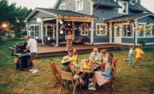 A group of people gathered around a table in the backyard of a blue house with a man working a grill nearby.