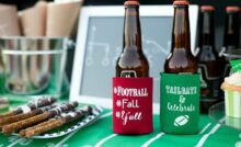 Two glass beer bottles with koozies, cupcakes, and chocolate-dipped pretzels sit on a table with football-themed decorations.